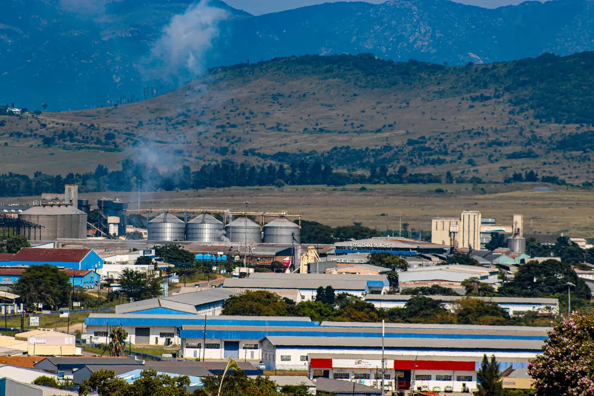 A panoramic view of an industrial area with silos, smokestack, and distant mountains under a clear blue sky.