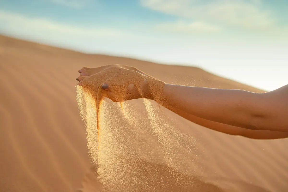 A hand releasing fine sand against the vast landscape of Merzouga Sand Dunes, Morocco.